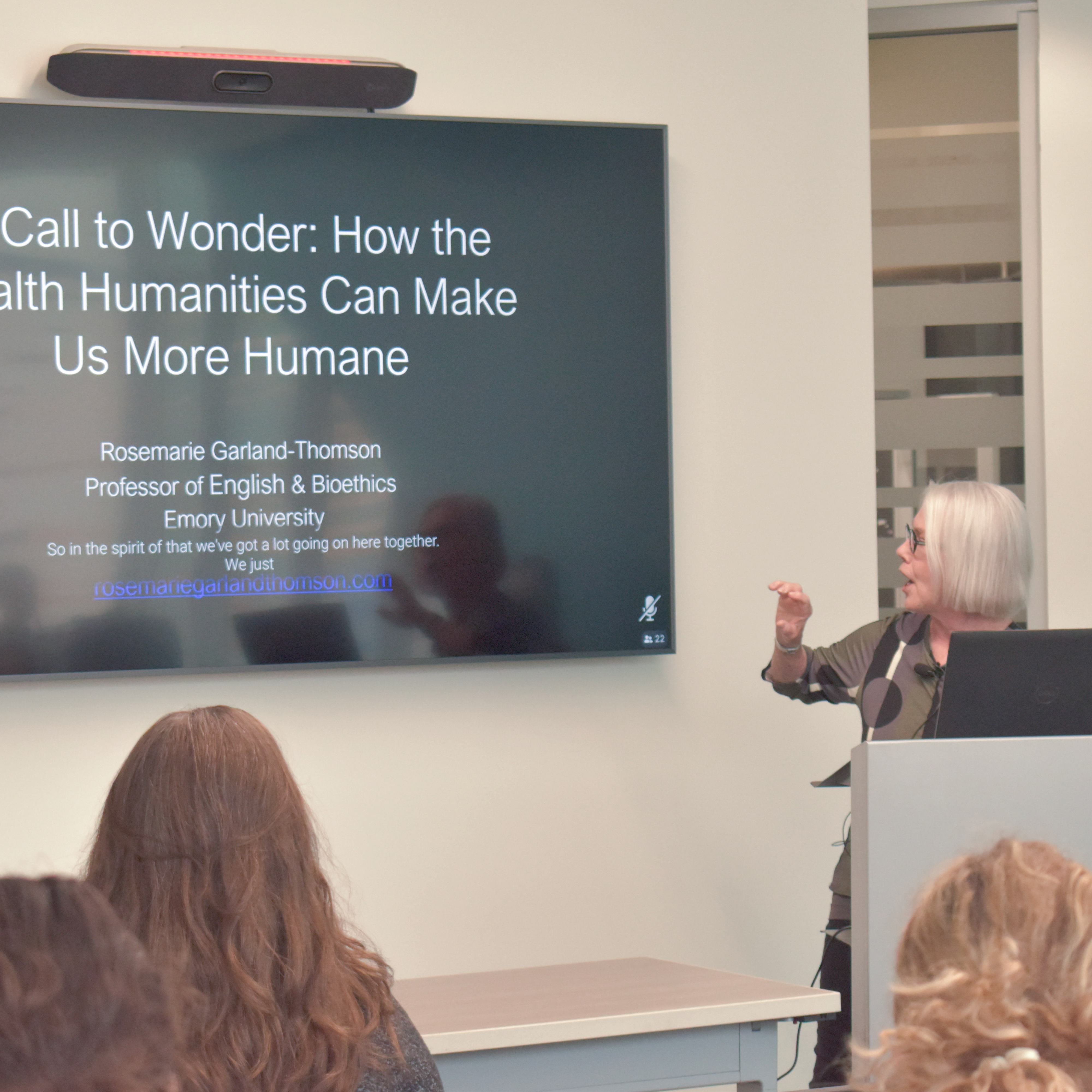 A woman with short, silvery hair stands at a podium, turned toward the monitor as she delivers a presentation.