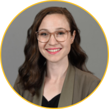 Woman with dark, wavy hair and pale skin wearing glasses and a brown suit jacket